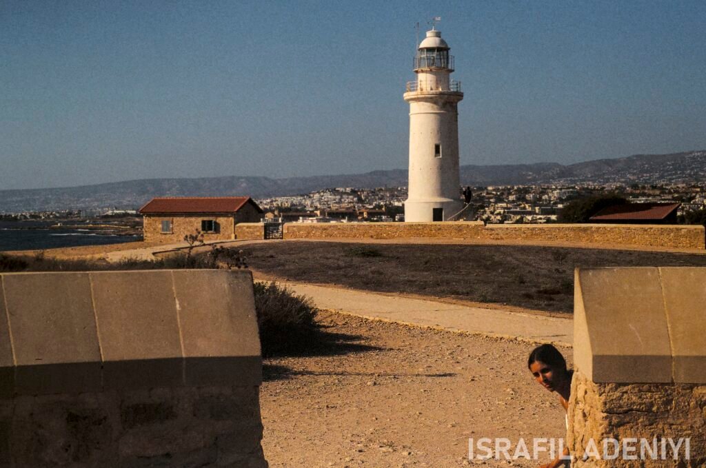 Lady peeking around a wall. Lighthouse in the distance, with shack beside.